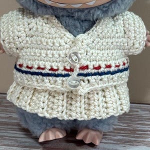 May include: A cream-colored, crocheted sweater set with a V-neck cardigan and a skirt. The sweater features red and blue stripes and two clear buttons. The set is designed for a plush toy.