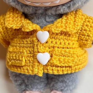 May include: A handmade yellow crochet jacket with white heart-shaped buttons. The jacket is worn by a gray plush toy. The jacket has a collar and pockets. The toy has a smiling face.