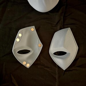 May include: Two gray plastic masks with a cutout for the eyes and nose. The masks have small, round, gold-colored magnets attached to the edges.