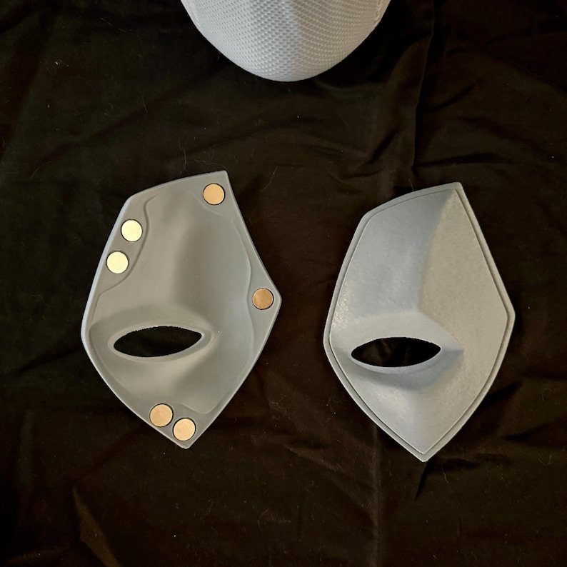 May include: Two gray plastic masks with a cutout for the eyes and nose. The masks have small, round, gold-colored magnets attached to the edges.