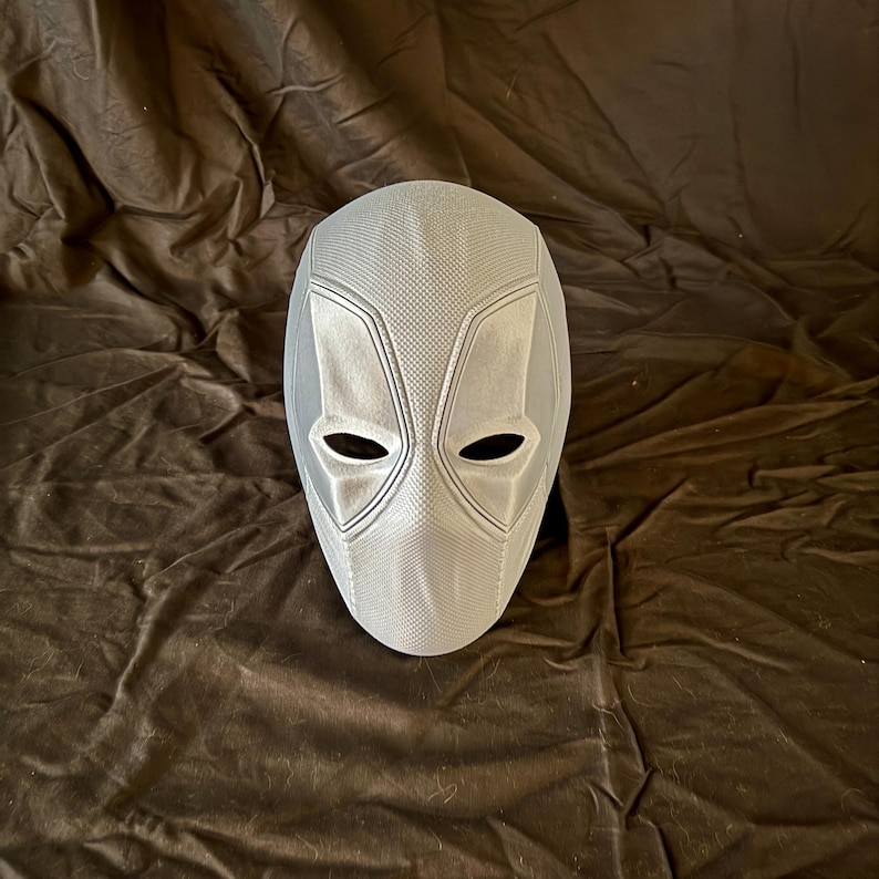 May include: A white plastic Deadpool mask with a textured surface. The mask has a black eye hole design.