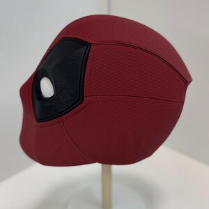 May include: A red and black Deadpool mask with a white eye hole. The mask is made of a textured material and has a detailed design.