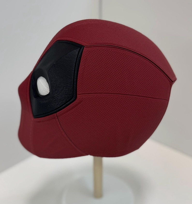 May include: A red and black Deadpool mask with a white eye hole. The mask is made of a textured material and has a detailed design.