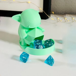 Puede incluir: Un soporte para dados verde menta con forma de criatura estilizada, que sostiene una forma geométrica. Varios dados azul claro con números blancos están dispersos alrededor del soporte y dentro de la bandeja.