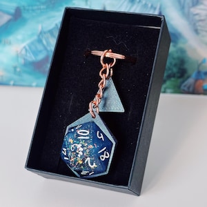 Può includere: Un portachiavi con dado D20 in resina blu e trasparente con catena e anello color rame. Il dado ha numeri bianchi e glitter colorati all'interno. Il portachiavi è in una scatola nera.