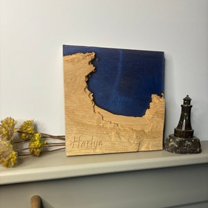 May include: A wooden wall art piece with a blue resin inlay depicting a coastline. The piece is personalised with the name "Hartlyn" in a cursive font.