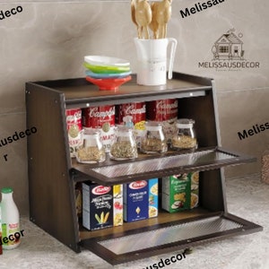 May include: A brown wooden cabinet with two shelves and glass doors. The top shelf has a variety of canned goods and jars of spices. The bottom shelf has boxes of pasta and other food items. The cabinet has a metal handle and is sitting on a tiled floor.