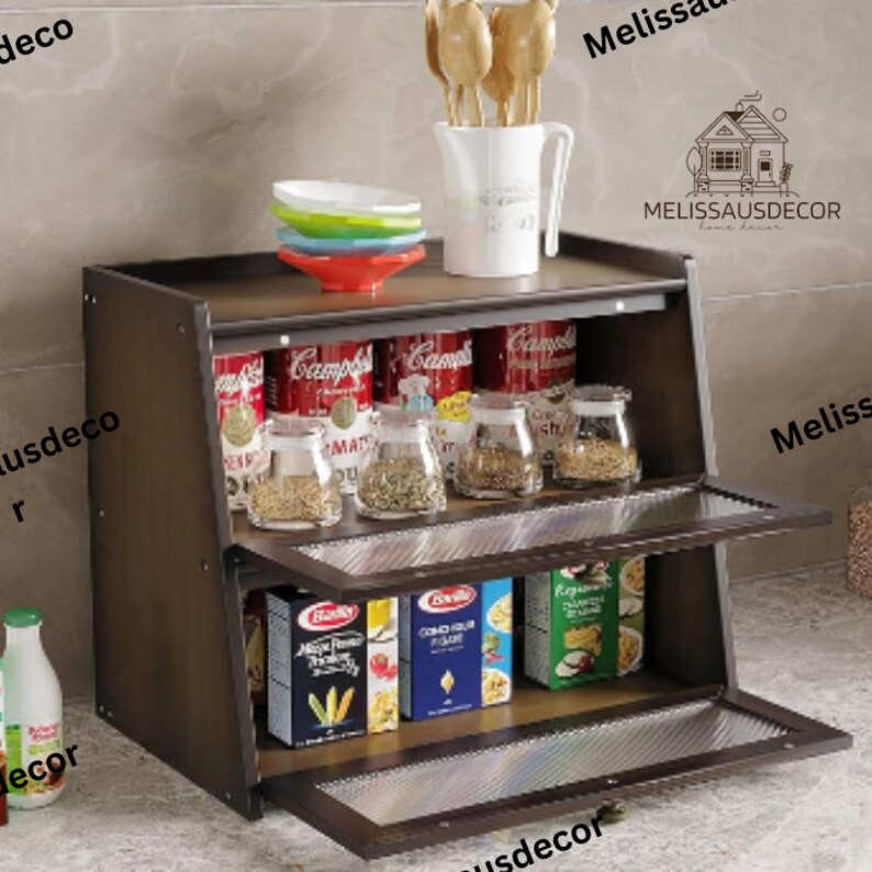 May include: A brown wooden cabinet with two shelves and glass doors. The top shelf has a variety of canned goods and jars of spices. The bottom shelf has boxes of pasta and other food items. The cabinet has a metal handle and is sitting on a tiled floor.