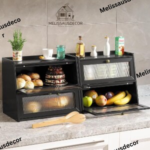May include: Black metal bread box with two shelves and glass doors. The top shelf has a cake and the bottom shelf has bread. The bread box is on a countertop with a wooden spatula.