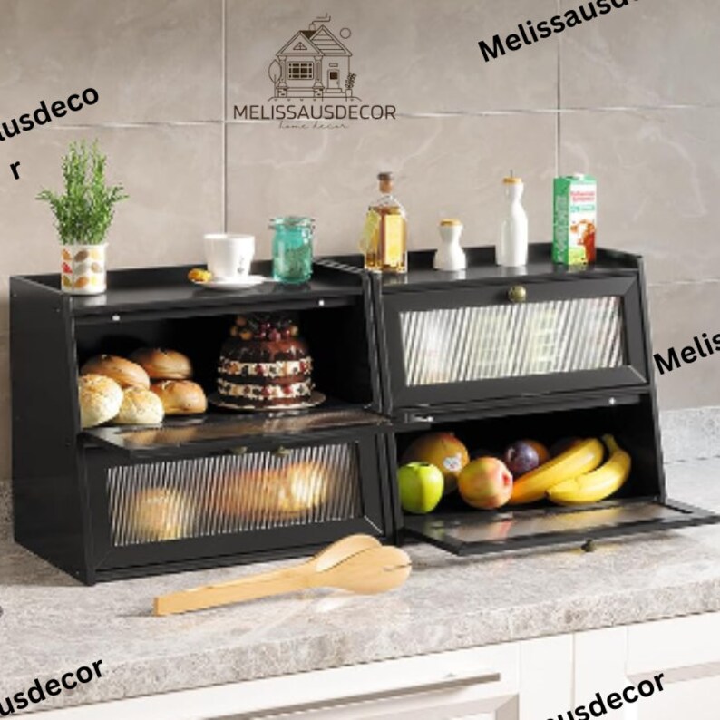 May include: Black metal bread box with two shelves and glass doors. The top shelf has a cake and the bottom shelf has bread. The bread box is on a countertop with a wooden spatula.