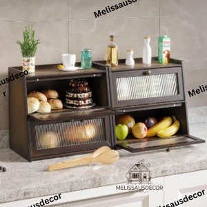 May include: A brown wooden bread box with three shelves and glass doors. The top shelf is empty, the middle shelf has a cake and bread, and the bottom shelf has fruit and bread. The bread box is on a countertop.