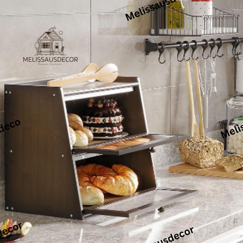 May include: A two-tiered brown wooden bread box with a glass door. The top tier has a cake and bread rolls inside. The bottom tier has croissants and bread rolls inside. The bread box is on a white countertop.