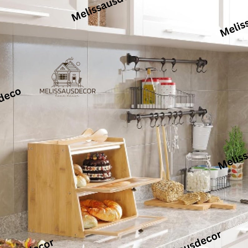 May include: A two-tiered bamboo bread box with a glass door and a wooden top. The bread box is filled with bread and pastries. The bread box is sitting on a countertop in a kitchen. There are hooks on the wall above the bread box.