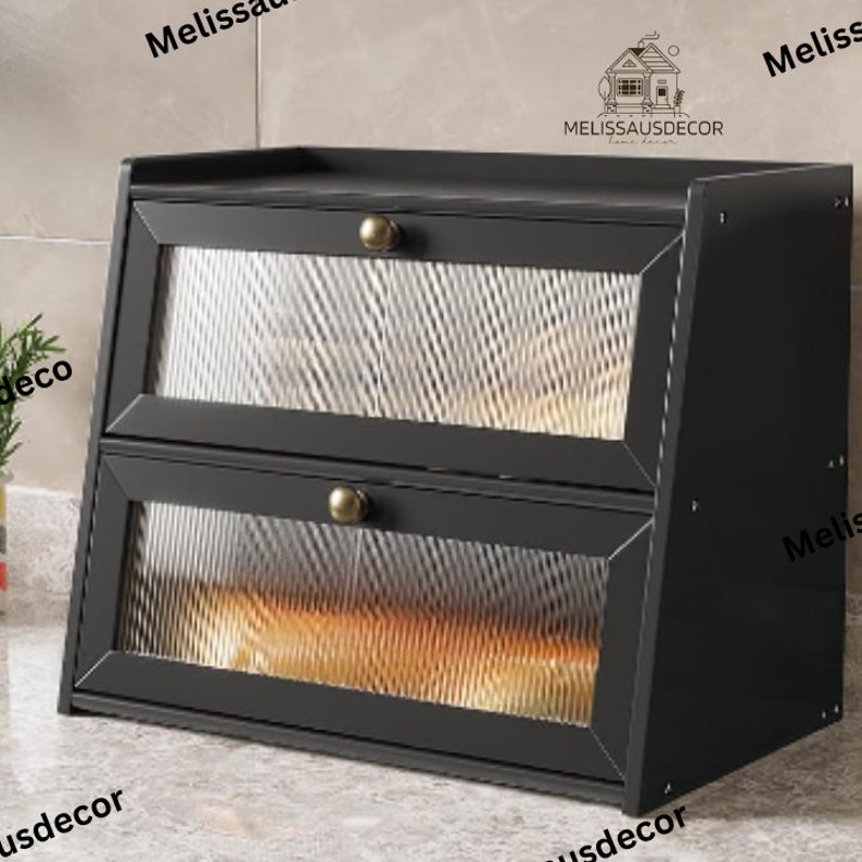 May include: Black two-tiered bread box with glass doors and gold-toned handles. The bread box has a decorative design on the glass doors.