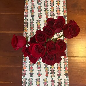 May include: A white table runner with a floral pattern of pink, yellow, and green roses. A bouquet of red roses is on top of the runner.