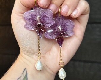 Könnte beinhalten: Ein Paar Ohrringe mit in Harz konservierten violetten Orchideenblütenblättern, die an goldfarbenen Creolen befestigt sind. Jeder Ohrring hat eine zarte Goldkette, die von der Blüte herabhängt und in einer kleinen, weißen, unregelmäßig geformten Perle endet.