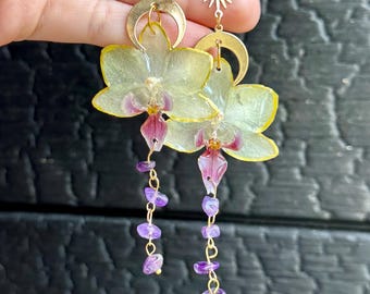 May include: A pair of gold-tone dangle earrings featuring a star and sun charm, a crescent moon, a translucent pale yellow orchid flower with a purple center, and a dangling chain of small, irregular purple amethyst gemstone beads.