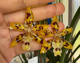 May include: A pair of hoop earrings featuring preserved yellow orchid flowers with dark red spots. The petals have a glossy, resin-coated finish, and each flower is attached to a thin, circular gold-toned metal hoop wire for hanging.