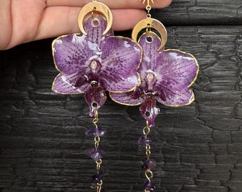 May include: A pair of dangle earrings featuring preserved purple orchid petals with gold-toned edges. Each earring includes a gold-toned crescent moon charm, one with a star and one with a sun, and a dangling chain of small purple amethyst stone chips.