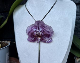 Pode incluir: Um colar com um pendente de uma flor de orquídea realista em tons de roxo e branco com veios roxos escuros. A orquídea está presa a uma fina corrente de cobra em tom prateado metálico que pende verticalmente do centro da flor.