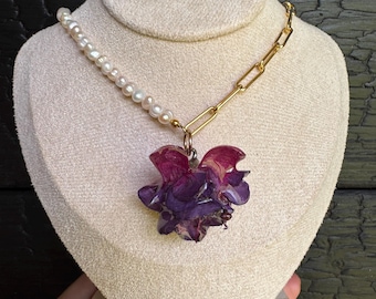Pode incluir: Um colar com metade de uma corrente de pequenas pérolas brancas e metade de elos de metal dourado em forma de clipe. Um grande pingente de pétalas múltiplas em tons de roxo profundo e magenta pende do centro da corrente.