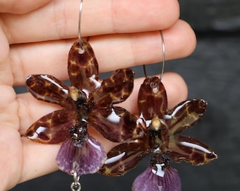 May include: A pair of hoop earrings featuring glossy, brown and tan mottled orchid-shaped pendants with purple lower petals. Each earring is accented with a small, dangling, teardrop-shaped purple crystal charm suspended from the flower base.