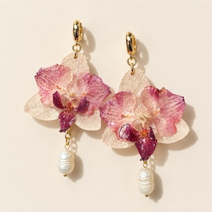 Puede incluir: Un par de pendientes de aro dorados con flores de orquídea prensadas de pétalos rosas y morados oscuros. Cada pendiente está adornado con una perla de agua dulce blanca de forma ovalada que cuelga de la parte inferior.