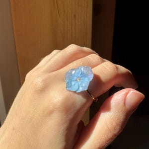 May include: A ring featuring a single, light blue, pressed hydrangea flower petal encased in a clear, glossy resin dome. The flower is attached to a thin, metallic band that encircles the finger, showcasing a delicate and botanical design.