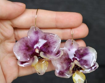 May include: A pair of dangle earrings featuring preserved orchid flowers with deep purple and white petals and a yellow center. The flowers are coated in a glossy, clear resin finish and are attached to thin, gold-toned metal earring hooks.