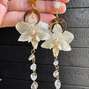 May include: A pair of dangle earrings featuring preserved white orchid flowers, gold-tone crescent moon charms, and a gold star or sun charm. Each earring includes a gold-tone hook and a vertical chain of small, clear, irregular crystal-like beads.