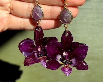 May include: Dangle earrings featuring a teardrop-shaped purple stone set in gold-toned metal, connected to a large, glossy, dark purple orchid flower pendant with intricate petal details and a gold-toned hook earring finding.