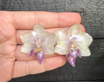 May include: A pair of earrings featuring preserved orchid flowers encased in clear resin. The orchids have pale cream petals with delicate purple centers and small yellow details, showcasing a natural floral shape and glossy finish.