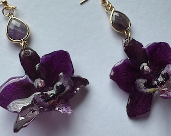 May include: A pair of dangle earrings featuring deep purple orchid petals preserved in a glossy resin finish. Each earring is attached to a gold-toned hook and includes a faceted, teardrop-shaped purple gemstone accent positioned above the flower.
