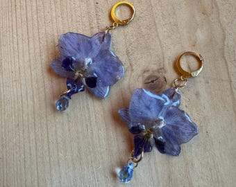May include: A pair of dangle earrings featuring preserved purple orchid petals encased in a clear resin finish. Each earring is attached to a gold-toned metal leverback clasp and includes a small, light blue teardrop-shaped crystal bead.