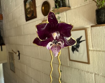 Pode incluir: Um gancho de cabelo com uma orquídea roxa escura preservada e uma borda metálica dourada. A flor está presa a um alfinete de metal dourado de duas pontas onduladas. As pétalas da orquídea apresentam um acabamento brilhante e salpicado.