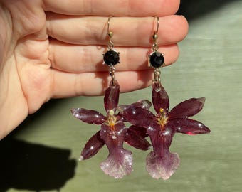 Pode incluir: Um par de brincos com uma flor de orquídea roxa escura e um pingente de pedra preciosa preta. Os brincos têm ganchos de ouro.