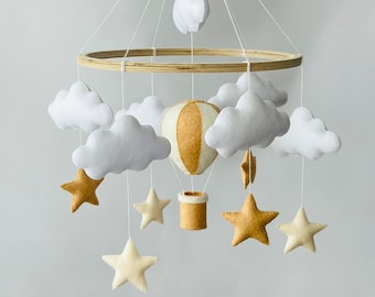 May include: A white and beige felt baby mobile with a hot air balloon, clouds, and stars hanging from a wooden ring.