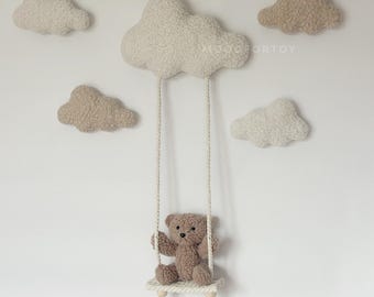 Könnte beinhalten: Ein brauner Teddybär sitzt auf einer Schaukel aus Seil und Holz. Die Schaukel hängt an einer großen, weißen, flauschigen Wolke. An der Wand hinter der Schaukel befinden sich vier kleinere, weiße und beige, flauschige Wolken.