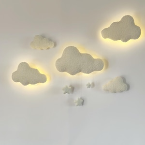 May include: A set of cloud-shaped wall lights and star-shaped hanging decorations. The lights are a soft white color with a warm yellow glow emanating from behind them. The stars are a matching white color.
