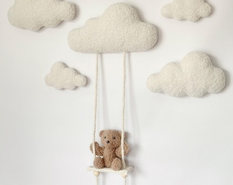 May include: A nursery wall decor set featuring fluffy white cloud-shaped pillows and a teddy bear on a swing. The bear is brown and the swing is suspended from a larger cloud. The clouds are arranged on a white wall.