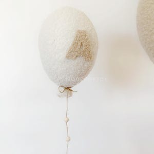 K&ouml;nnte beinhalten: Ein wei&szlig;er, flauschiger Ballon mit einem braunen Filzbuchstaben "A" daran befestigt. Der Ballon ist an einer Schnur befestigt und hat eine Schnur mit wei&szlig;en Holzperlen, die daran h&auml;ngen.