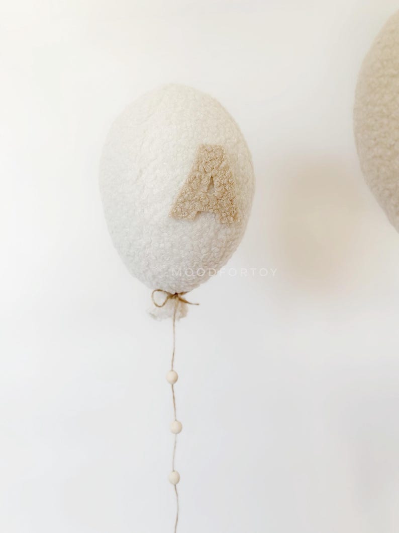 K&ouml;nnte beinhalten: Ein wei&szlig;er, flauschiger Ballon mit einem braunen Filzbuchstaben "A" daran befestigt. Der Ballon ist an einer Schnur befestigt und hat eine Schnur mit wei&szlig;en Holzperlen, die daran h&auml;ngen.