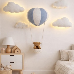 Op de afbeelding: Muurdecoratie voor de kinderkamer met een blauw-witte luchtballon en een teddybeer in een gevlochten mand. De muur is versierd met verlichte wolken. Een nachtkastje met een lamp en een mand met knuffels is ook zichtbaar.