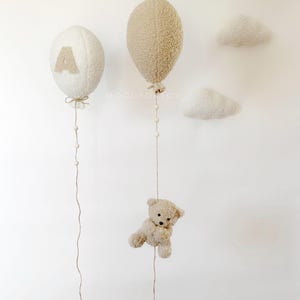 K&ouml;nnte beinhalten: Zwei pl&uuml;schige, cremefarbene Ballons mit Schn&uuml;ren, die von ihnen herabh&auml;ngen. Ein Ballon hat den Buchstaben 'A' darauf. Ein kleiner, cremefarbener Teddyb&auml;r h&auml;ngt an einer der Schn&uuml;re. Zwei cremefarbene Wolkenformen sind an der Wand befestigt.
