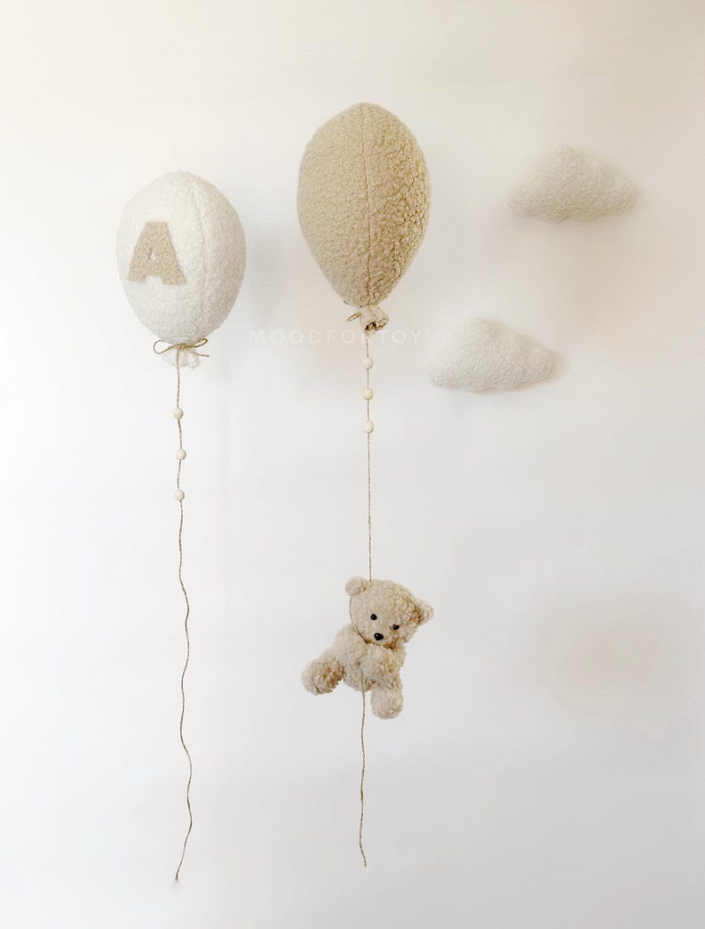 K&ouml;nnte beinhalten: Zwei pl&uuml;schige, cremefarbene Ballons mit Schn&uuml;ren, die von ihnen herabh&auml;ngen. Ein Ballon hat den Buchstaben 'A' darauf. Ein kleiner, cremefarbener Teddyb&auml;r h&auml;ngt an einer der Schn&uuml;re. Zwei cremefarbene Wolkenformen sind an der Wand befestigt.