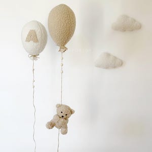 K&ouml;nnte beinhalten: Zwei flauschige, cremefarbene Ballons mit einem braunen Teddyb&auml;ren, der an einer der Schn&uuml;re h&auml;ngt. Die Ballons sind mit einem kleinen, braunen Filzberg und dem Buchstaben "A" verziert. Zwei wei&szlig;e, flauschige Wolken sind an der Wand befestigt.