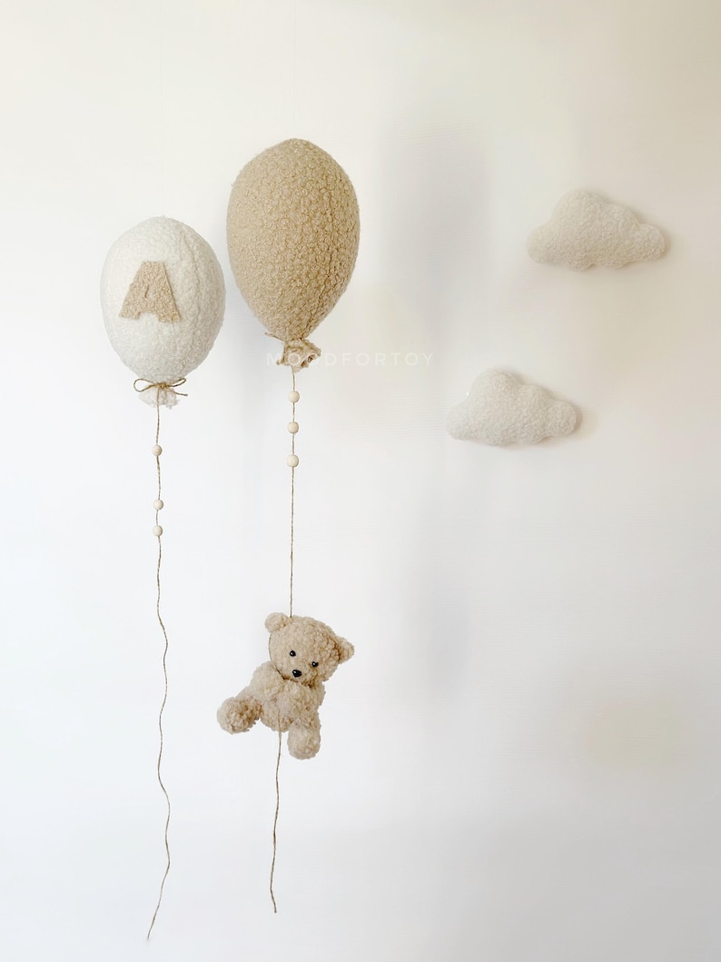 K&ouml;nnte beinhalten: Zwei flauschige, cremefarbene Ballons mit einem braunen Teddyb&auml;ren, der an einer der Schn&uuml;re h&auml;ngt. Die Ballons sind mit einem kleinen, braunen Filzberg und dem Buchstaben "A" verziert. Zwei wei&szlig;e, flauschige Wolken sind an der Wand befestigt.