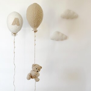 K&ouml;nnte beinhalten: Zwei flauschige wei&szlig;e und braune Ballons mit Schnur und Perlen, die von ihnen h&auml;ngen. Ein kleiner brauner Teddyb&auml;r h&auml;ngt an der Schnur eines der Ballons. Zwei flauschige wei&szlig;e Wolken sind an der Wand befestigt.