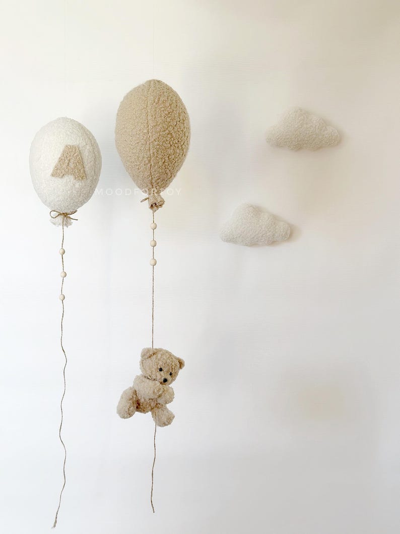 K&ouml;nnte beinhalten: Zwei flauschige wei&szlig;e und braune Ballons mit Schnur und Perlen, die von ihnen h&auml;ngen. Ein kleiner brauner Teddyb&auml;r h&auml;ngt an der Schnur eines der Ballons. Zwei flauschige wei&szlig;e Wolken sind an der Wand befestigt.