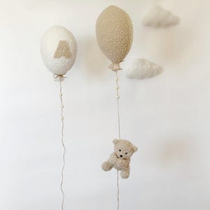 K&ouml;nnte beinhalten: Zwei flauschige, cremefarbene Ballons mit einem braunen Buchstaben 'A' auf einem Ballon und einem braunen Teddyb&auml;r, der am Boden des anderen Ballons h&auml;ngt. Die Ballons h&auml;ngen an einer wei&szlig;en Wand mit zwei wei&szlig;en, flauschigen Wolken.